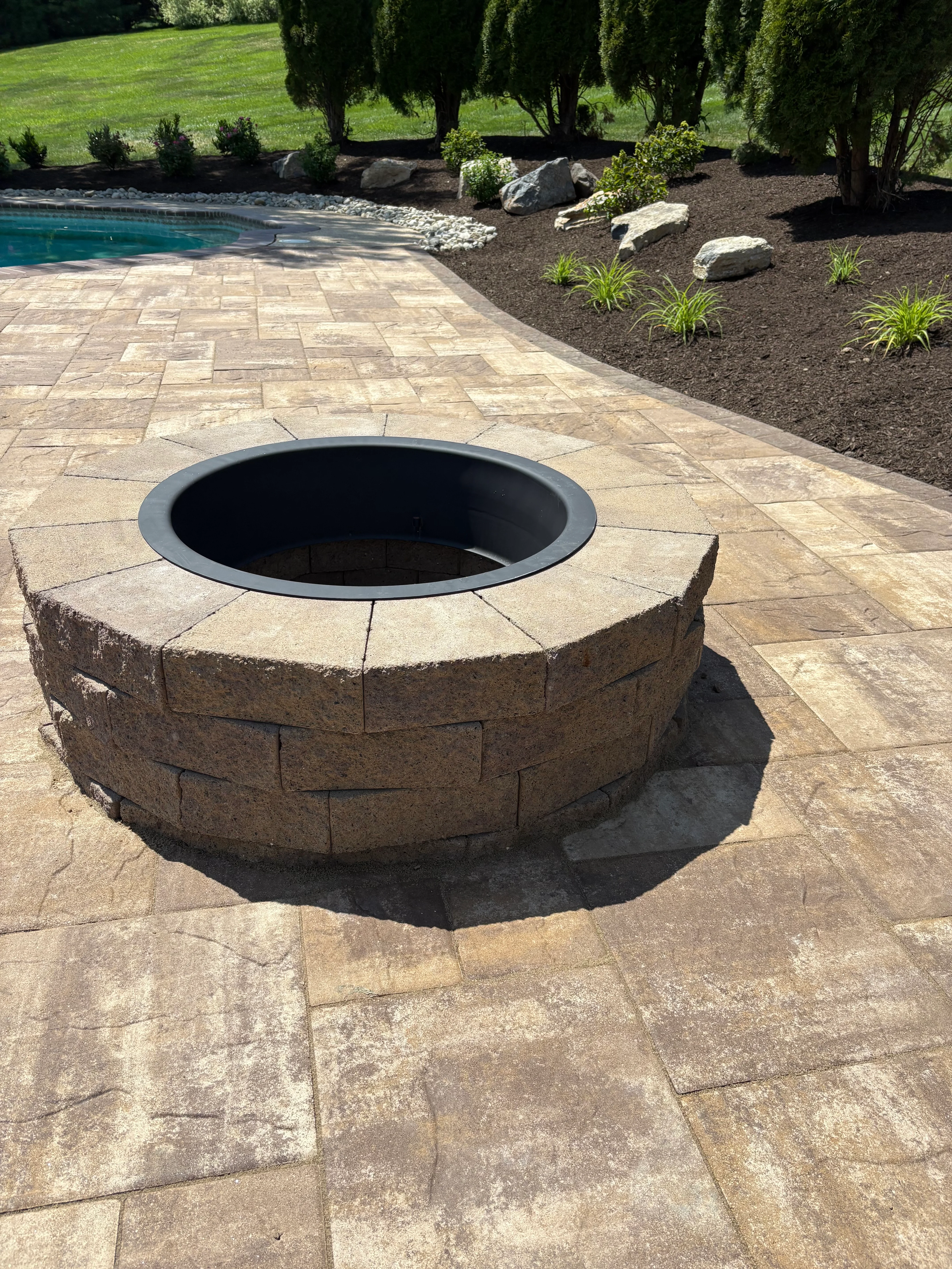 Stone fire pit with steel ring on stamped concrete patio beside pool and landscaped beds